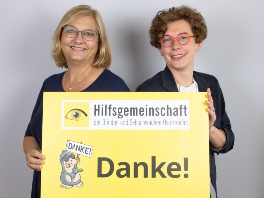 Zwei Mitarbeiterinnen der Hilfsgemeinschaft lächeln und halten ein gelbes Schild hoch, auf dem groß Danke steht
