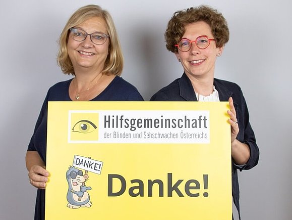 Zwei Mitarbeiterinnen der Hilfsgemeinschaft lächeln und halten ein gelbes Schild hoch, auf dem groß Danke steht