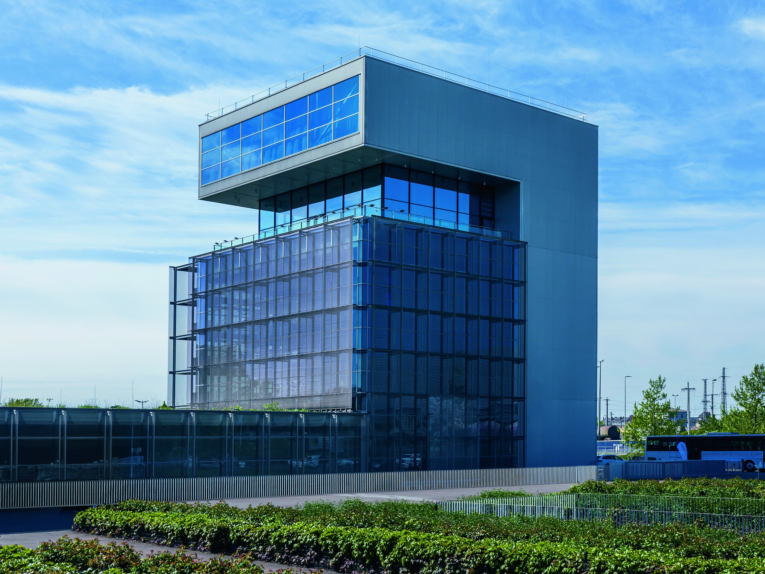Modernes, kubisches Gebäude mit großer Glasfassade und auskragendem Obergeschoss unter blauem Himmel; im Vordergrund begrüntes Gelände und ein Linienbus.