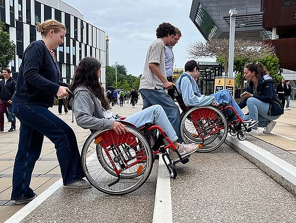 Jungen Menschen, die in Rollstühlen sitzen, wird von anderen jungen Menschen rüclwärts breite Stufen hinunter geholfen. Eine Frau kniet daneben und erklärt, wie es geht. 