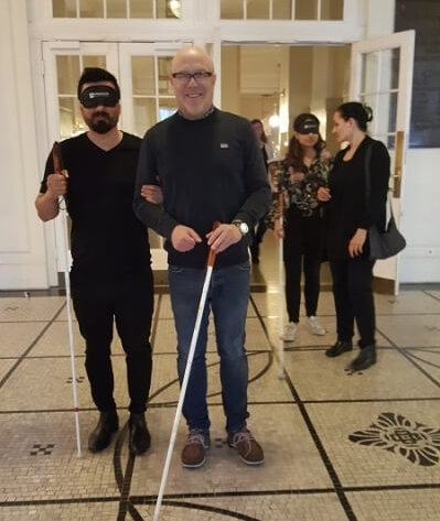 In einem Flur stehen ein Mann mit Augenbinde und ein sehbehindertes Mitglied, beide mit Langstock ausgerüstet, nebeneinander.