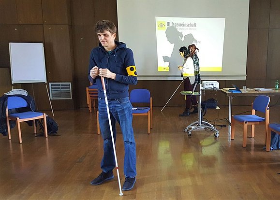 Mann mit weißem Stock und gelber Armbinde inmitten eines Sitzkreises. Hinter ihm ein Beamer mit Leinwand und zwei Personen wandernd mit Augenbinde und weißem Stock.