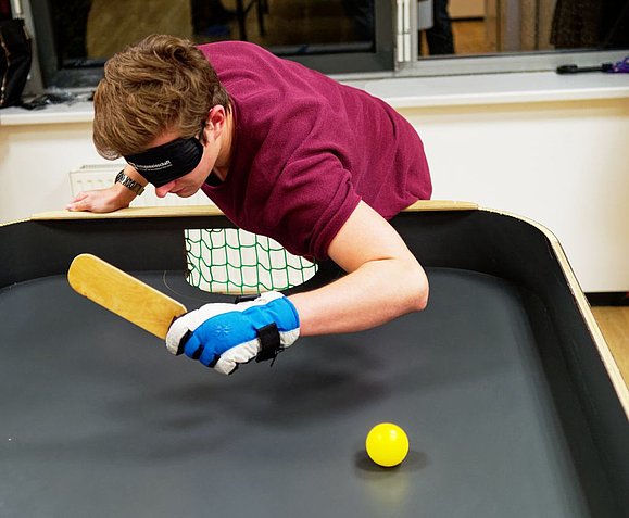 Junger Mann mit purpur T-Shirt, Handschuhen und Augenbinde spielt Tennisball auf Tisch mit länglichem Schläger