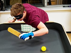 Junger Mann mit purpur T-Shirt, Handschuhen und Augenbinde spielt Tennisball auf Tisch mit länglichem Schläger
