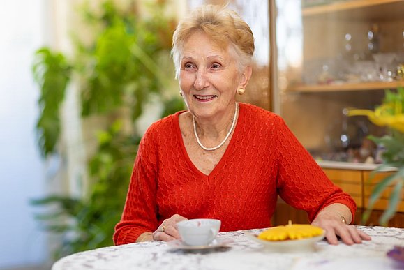 ältere Frau mit rotem Pullover und kurzen blonden Haaren sitzt am Tisch mit Kaffeetasse und lächelt