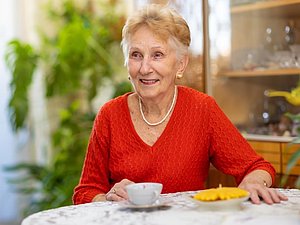 ältere Frau mit rotem Pullover und kurzen blonden Haaren sitzt am Tisch mit Kaffeetasse und lächelt