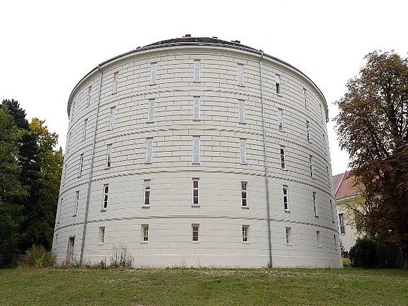 Weißer Turm mit vielen Fenster, Wiese und Bäume im Umkreis.