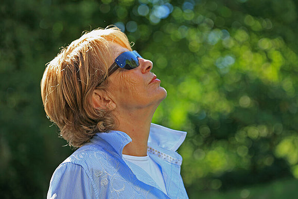Eine Frau mit Sonnenschutzbrille hat ihren Kopf in den Nacken gelegt und blickt in den Himmel, hinter ihr stehen Bäume 