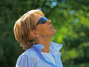Eine Frau mit Sonnenschutzbrille hat ihren Kopf in den Nacken gelegt und blickt in den Himmel, hinter ihr stehen Bäume 