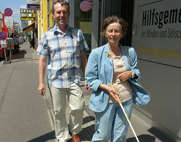 Mobilitätstrainer Milan Malecek begleitet ein weibliches Mitglied, das mit dem Langstock einem taktilen Bodenleitsystem folgt Sehbeeinträchtigtes weibliches Mitglied mit Langstock wird beim Mobilitätstraining begleitet