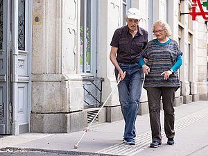 Frau mit grauen Haaren begleitet Mann mit Langstock am Gehsteig, Apothekenzeichen im Hintergrund 