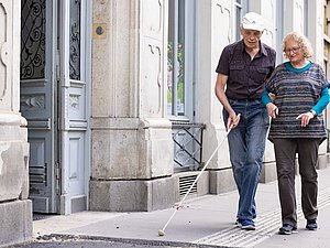 Frau mit grauen Haaren begleitet Mann mit Langstock am Gehsteig, Apothekenzeichen im Hintergrund 
