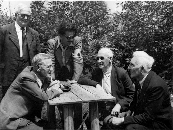  Zwei Männern stehen und drei Männer sitzen bei Tisch; Foto schwarz-weiß.