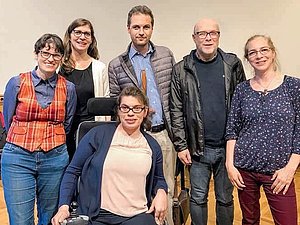 6 Menschen mit unterschiedlichen Behinderungen stehen gemeinsam in einem Saal mit braunem Boden.