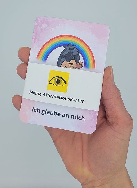 In einer Hand ist eine Karte mit dem Text "Ich glaube an mich"