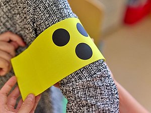 Eine Blindenschleife (drei schwarze Punkte auf gelben Hintergrund) wird auf einem Oberarm befestigt