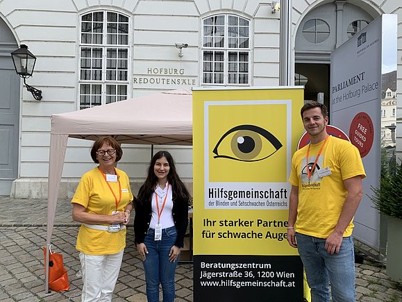 Ältere Frau mit Brille, junge Frau mit langen braunen Haaren und Junger Mann mit gelbem Shirt stehen neben gelbem Roll-Up der Hilfsgemeinschaft mit 