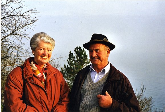 Ein älteres Pärchen steht nebeneinander in einer Herbstlandschaft, beide lächeln in die Kamera 