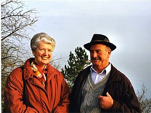 Ein älteres Pärchen steht nebeneinander in einer Herbstlandschaft, beide lächeln in die Kamera 