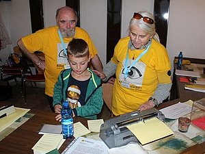 Ein Mann und eine Frau in gelben T-Shirts zeigen einem Jungen eine Braillemaschine. 
