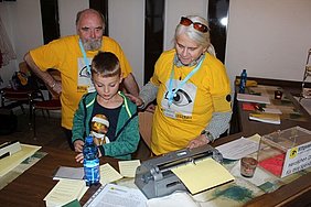Ein Mann und eine Frau in gelben T-Shirts zeigen einem Jungen eine Braillemaschine. 